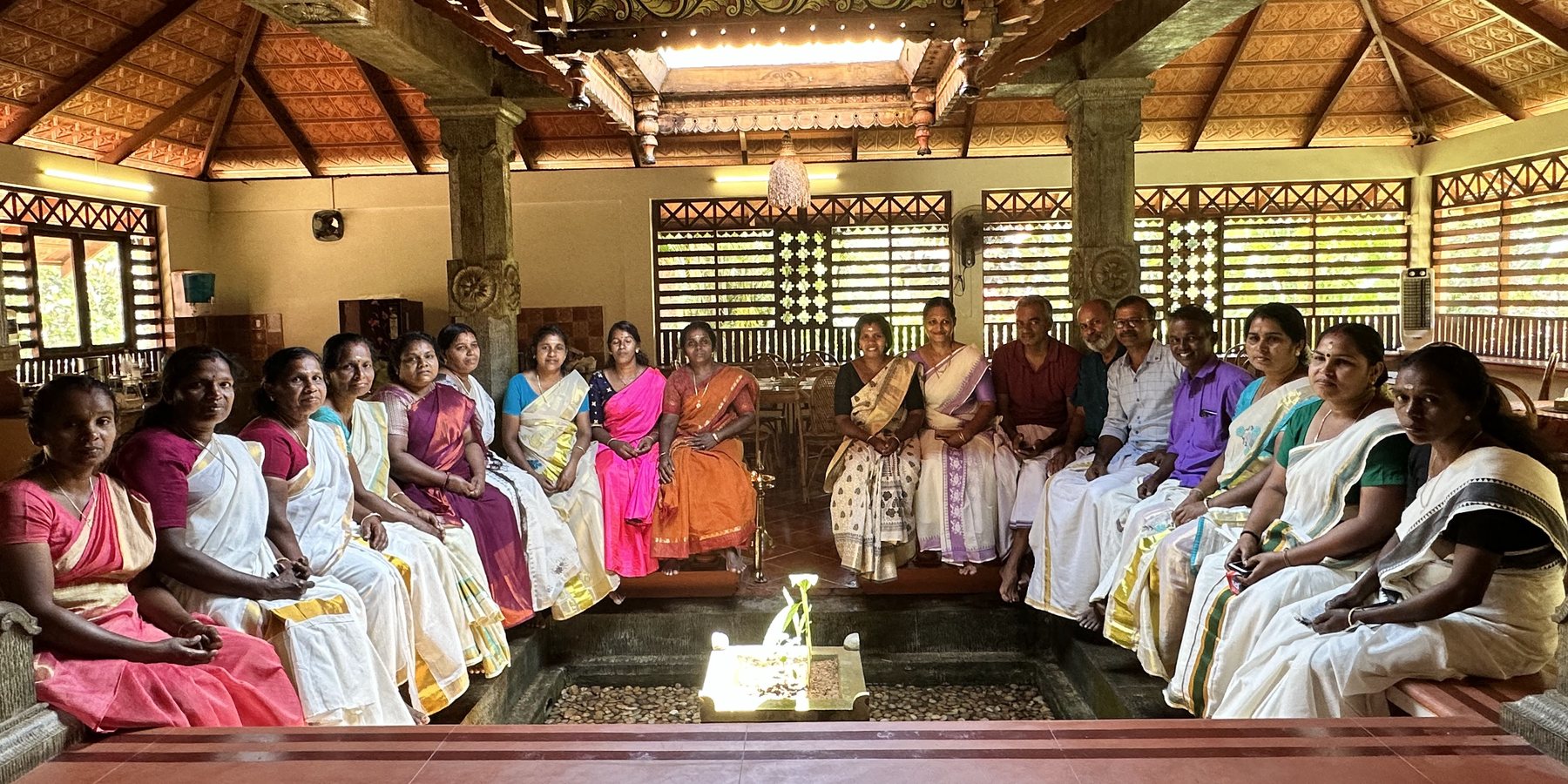 The Neelambari team seated around the Nadumuttam, the open-to-sky courtyard at the centre of the Nalukettu, under its carved timber roof - most of them women, in kasavu and coloured saris.
