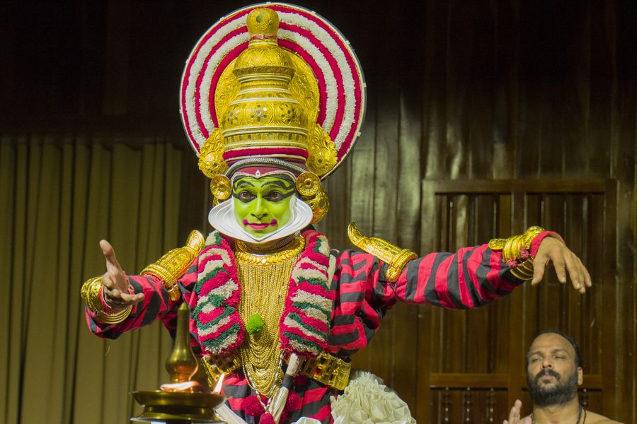 A Kathakali performer in full costume and makeup, arms outstretched beside a lit brass lamp.