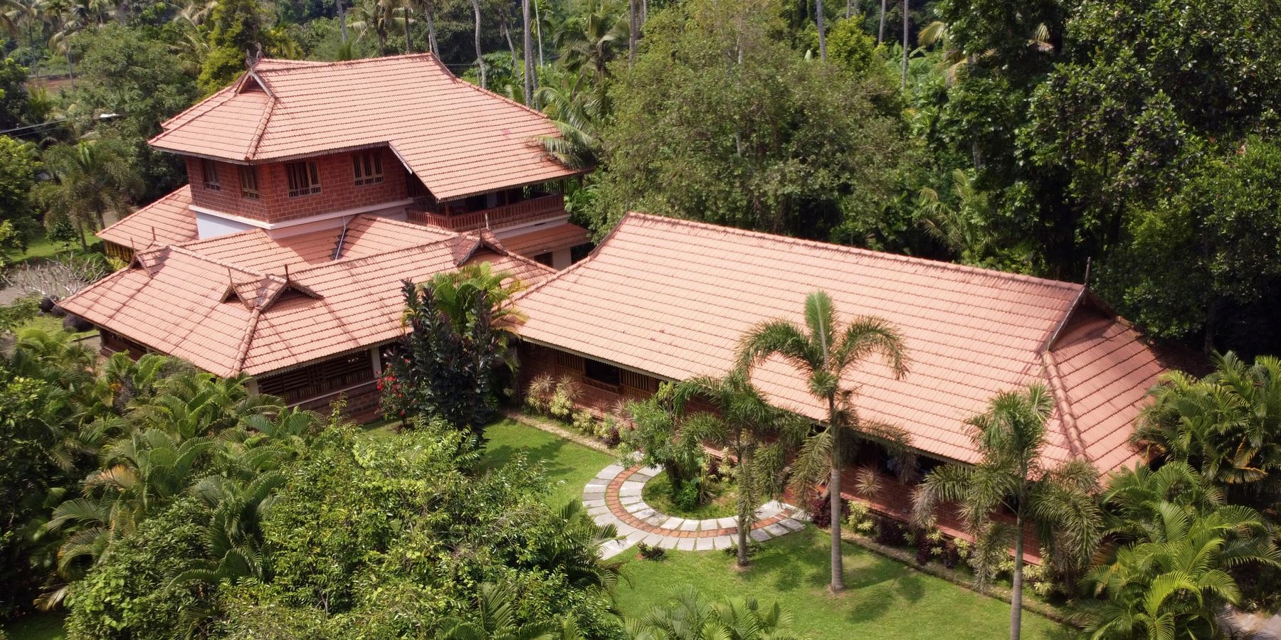 An aerial view of Neelambari's main building by day, its terracotta-tiled roofs set among coconut palms and garden, with a circular stone path leading to the entrance.