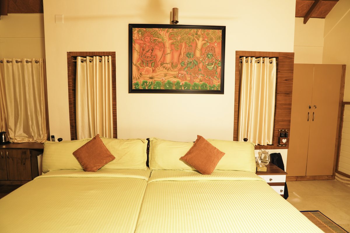 A deluxe room with a wide bed, orange bolsters, a framed Kerala mural above it and a timber wardrobe alongside.