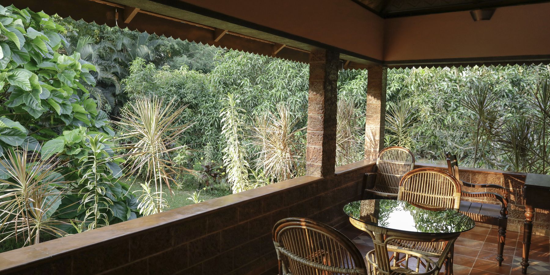 A room verandah at Neelambari with cane chairs and a low laterite wall, looking straight out into dense garden greenery.
