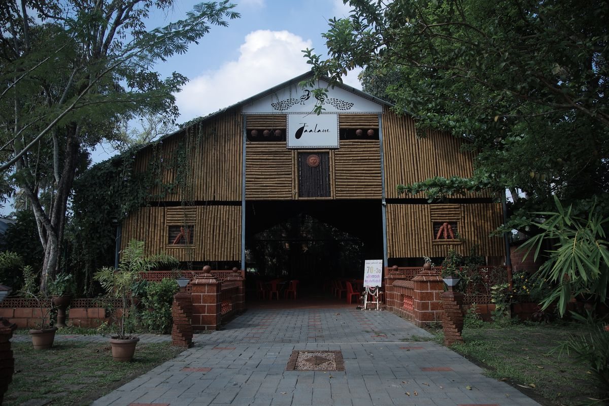 The bamboo frontage of Jaalam, creepers growing across it, with a paved path leading in through the open entrance to seating inside.