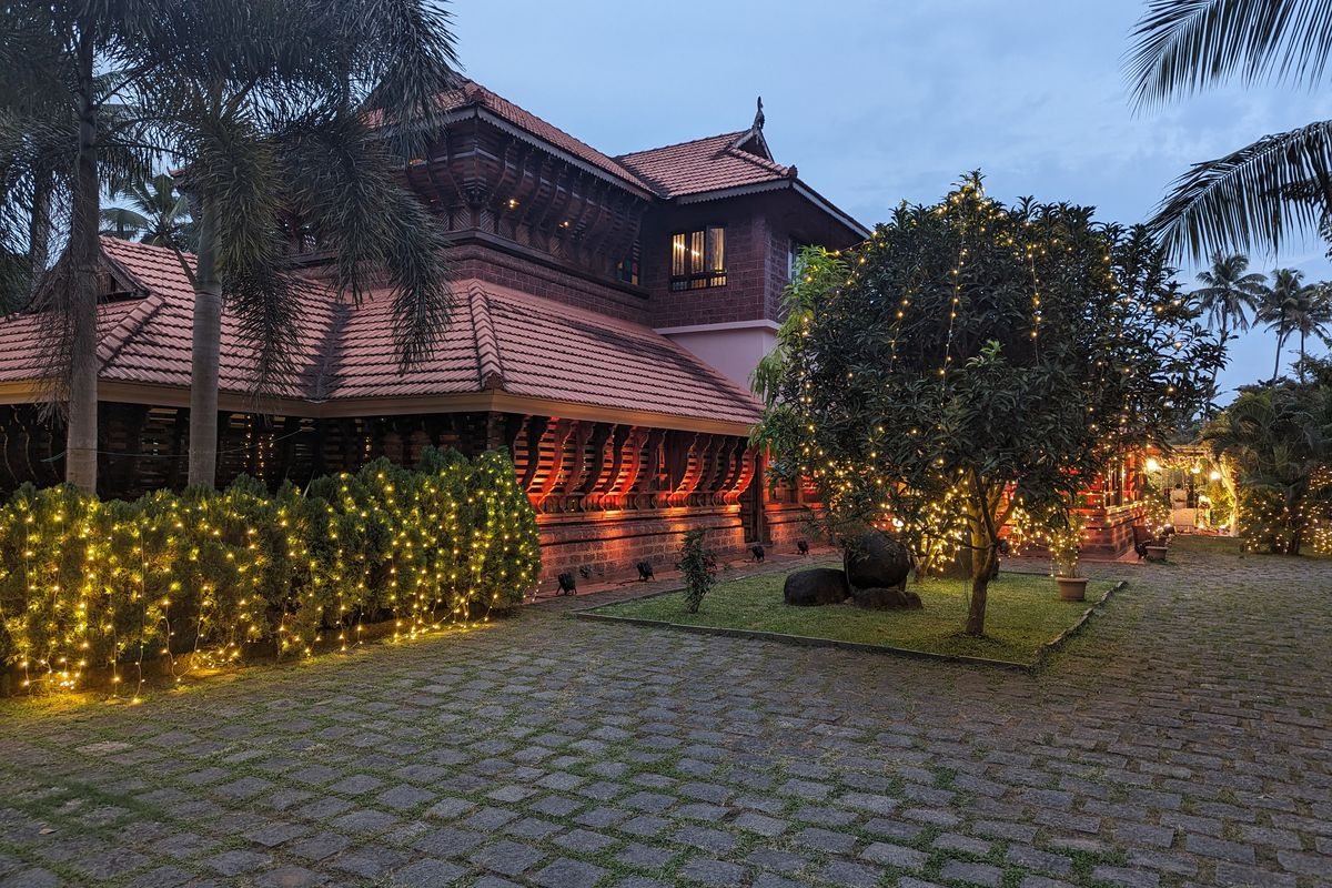The Koothambalam at dusk, its tiled roofs and carved verandah lit warm against a blue evening sky, with fairy lights strung through the hedge around the paved forecourt.
