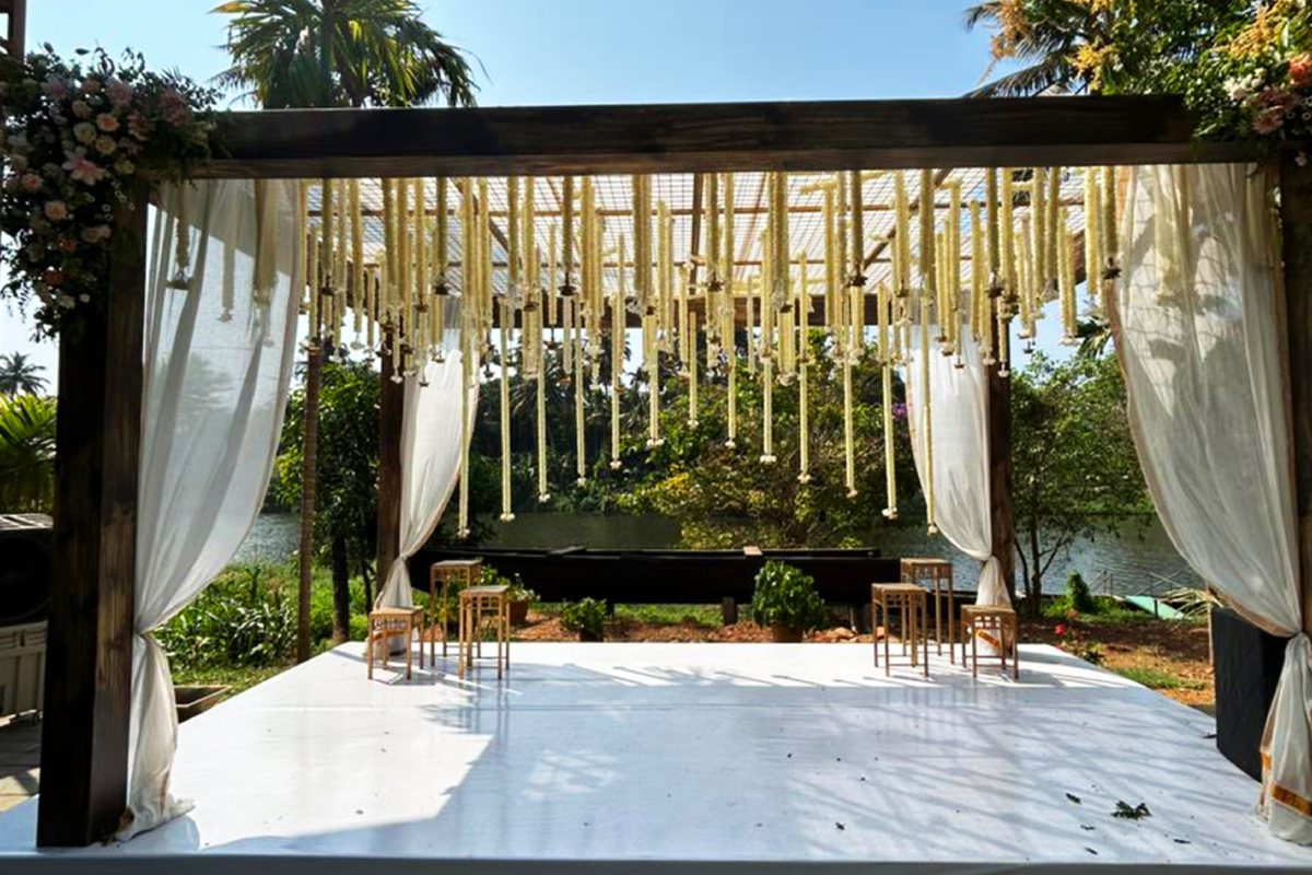 The riverside mandap at Puzhayoram, white drapes tied back at its timber posts and strings of jasmine hanging from the frame, looking through to the Karuvannur beyond.