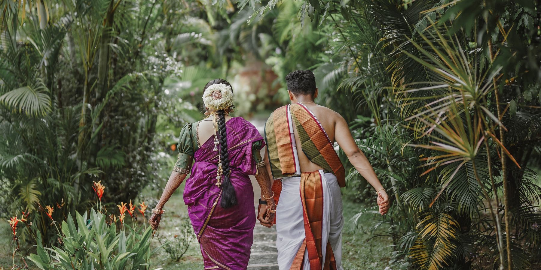 A bride and groom in traditional Kerala dress walking away hand in hand along a stone path at Neelambari, between dense palms and flowering garden.